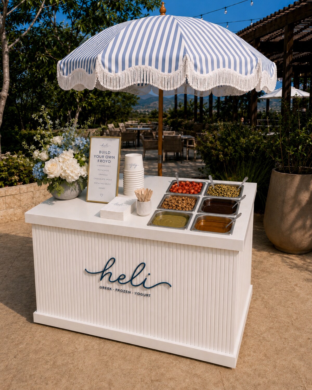 Heli Greek frozen yogurt event cart with a striped umbrella, branded counter, flowers, and topping bar